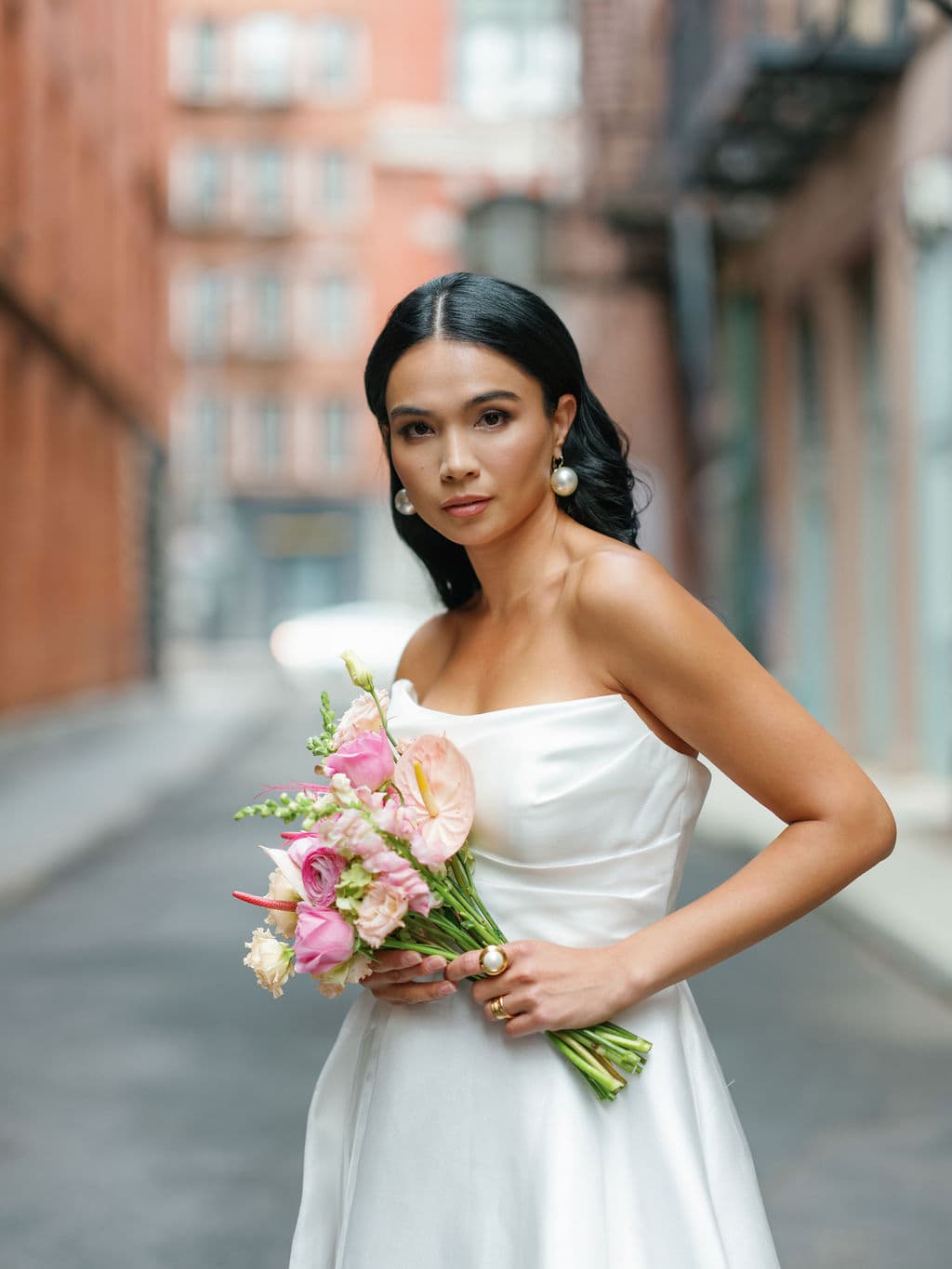Close-up of Shirin Hojayeva holding a pink floral bouquet in a white strapless dress on a brick-lined city street.