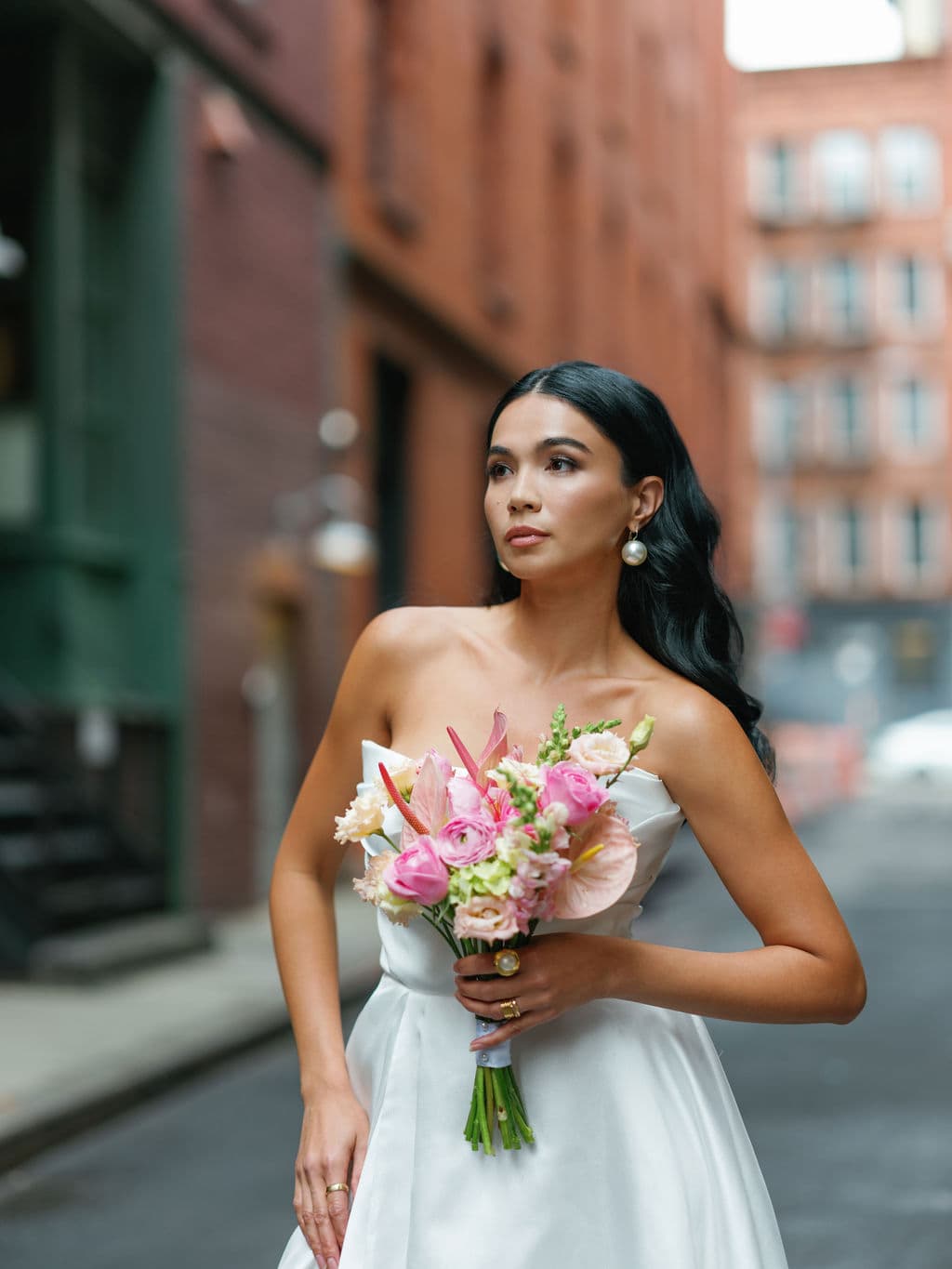 Shirin Hojayeva clutches a pink bouquet to her chest in a white strapless dress, looking away on an urban side street.