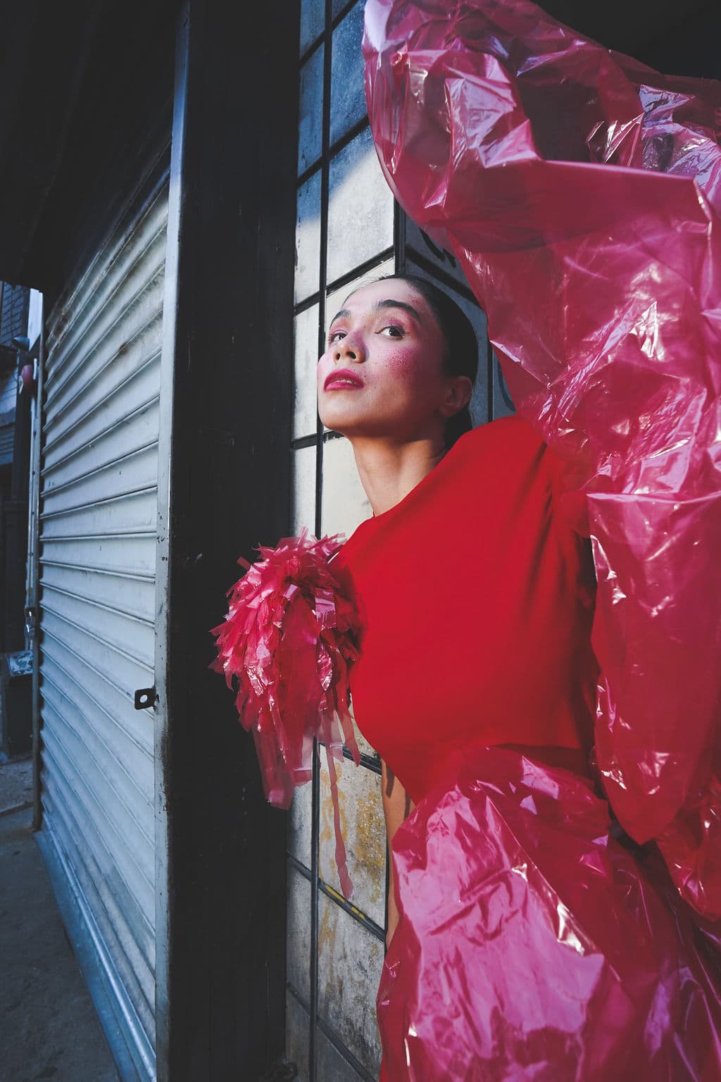 Shirin Hojayeva leans against a storefront in a red dress with pink cellophane ruffles, dramatic makeup, and bold styling.