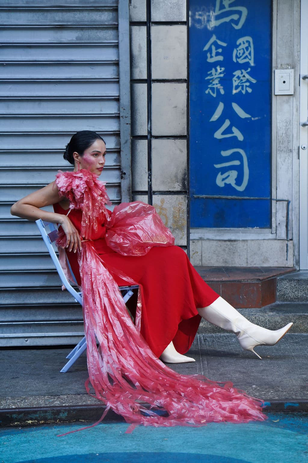 Shirin Hojayeva reclines on a chair in a red dress with pink plastic accents in front of a Chinatown shopfront.