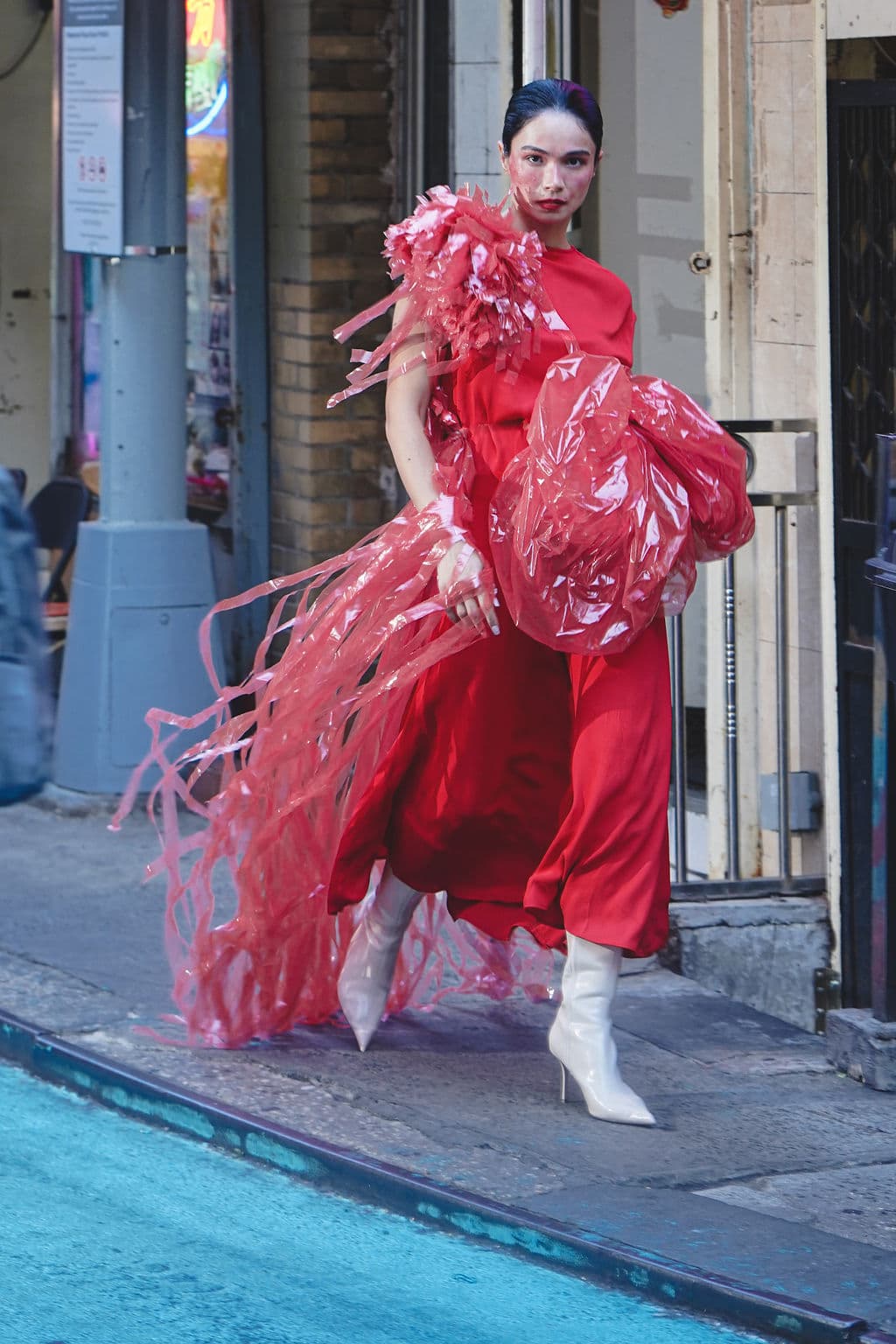 Shirin Hojayeva strides down a Chinatown sidewalk in a red dress with flowing pink plastic embellishments and white boots.