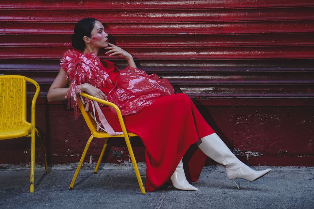 Shirin Hojayeva lounges on a yellow chair against a red metal shutter in a red dress with pink accents, gazing up.
