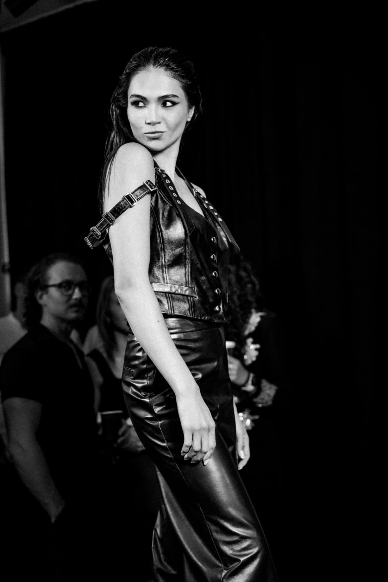 Black-and-white photo of Shirin Hojayeva backstage in a leather halter top and pants, glancing over her shoulder confidently.