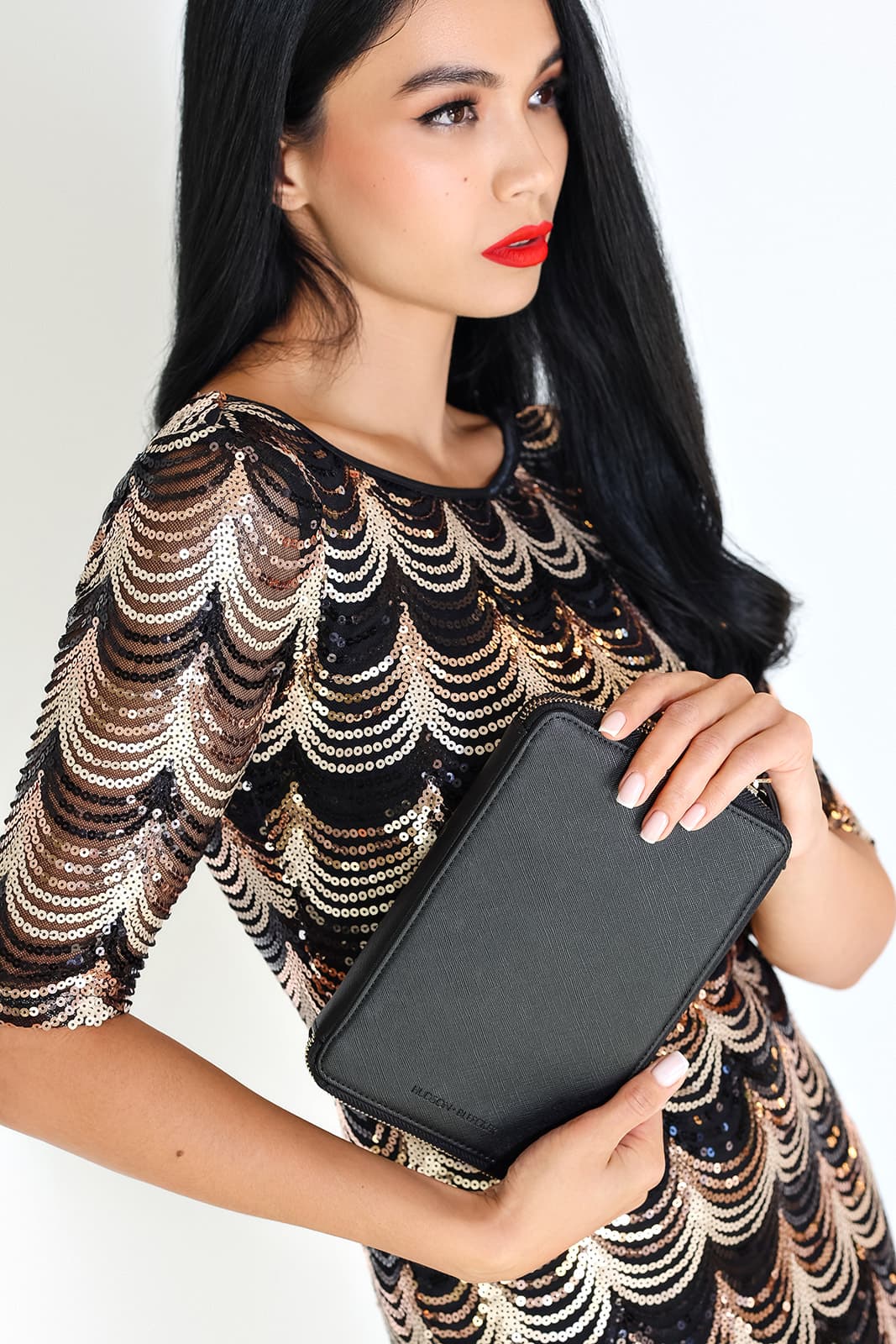 Shirin Hojayeva in a gold sequin scallop-pattern dress holds a black clutch, with red lips on a white backdrop.