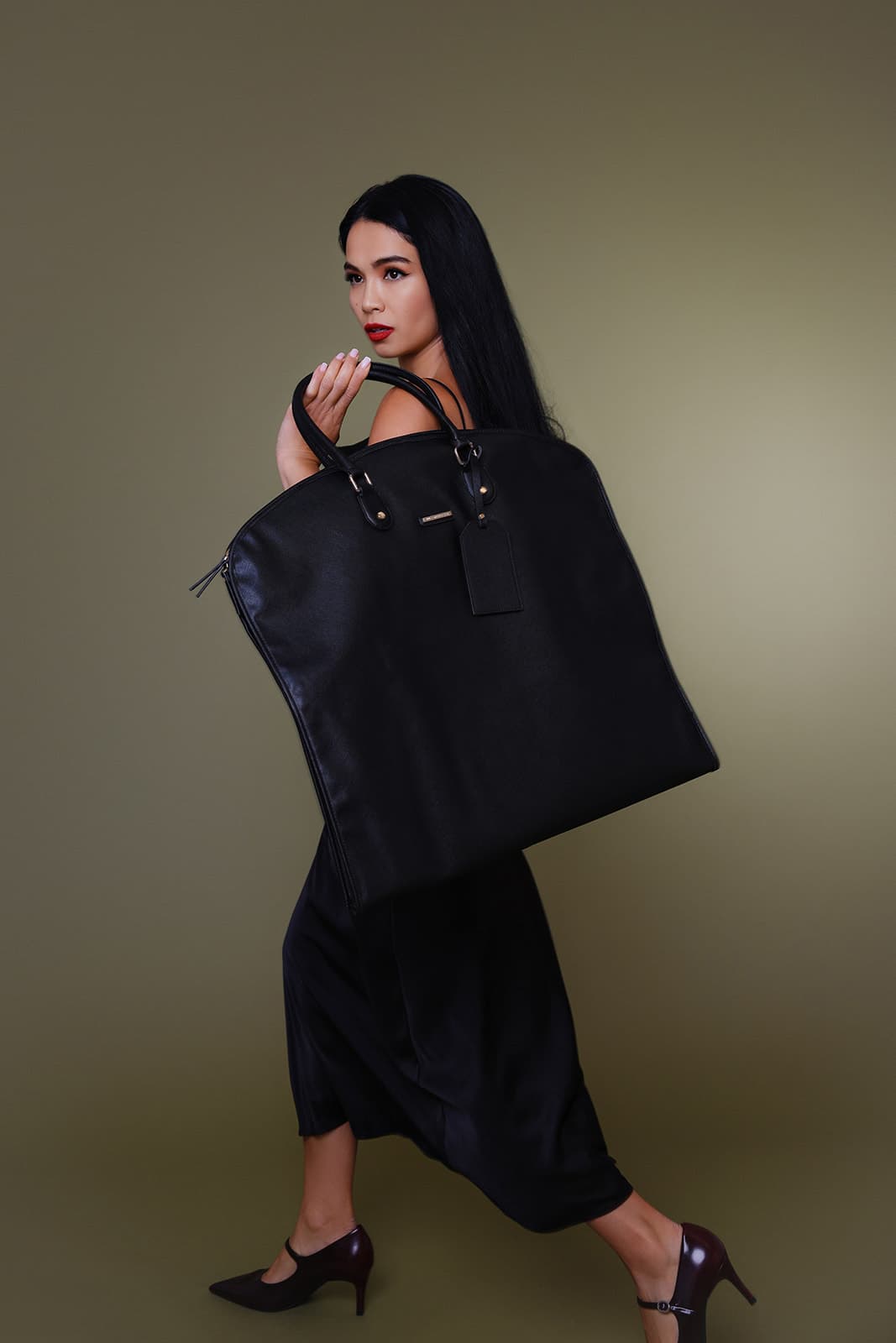 Shirin Hojayeva poses mid-stride in all black holding an oversized leather tote over her shoulder against an olive backdrop.