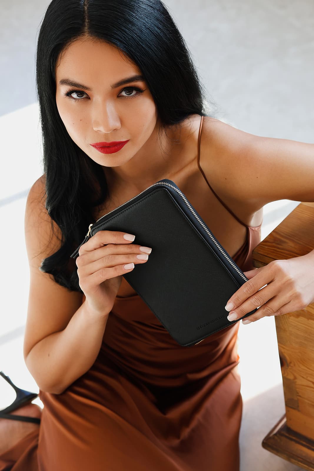 Shirin Hojayeva sits in a rust satin slip dress holding a black zippered wallet, looking up with bold red lips.