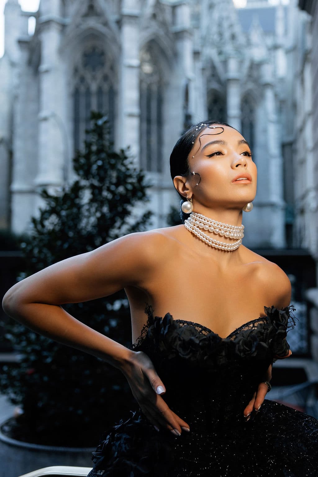 Close-up of Shirin Hojayeva in a black sparkle corset with pearl choker and earrings, cathedral spires in the background.